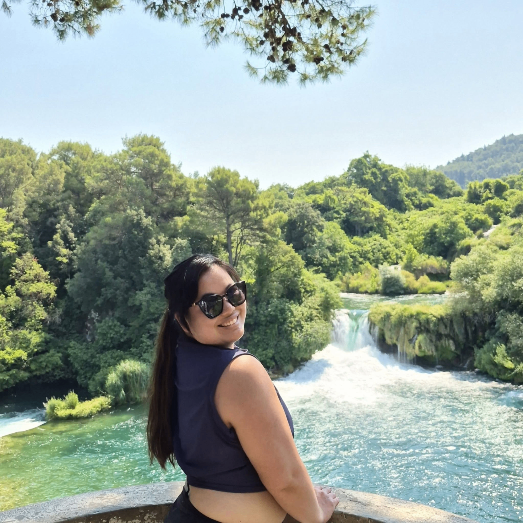 Katie of Wonderbound Travel smiling by a bright turquoise waterfall surrounded by lush green forest