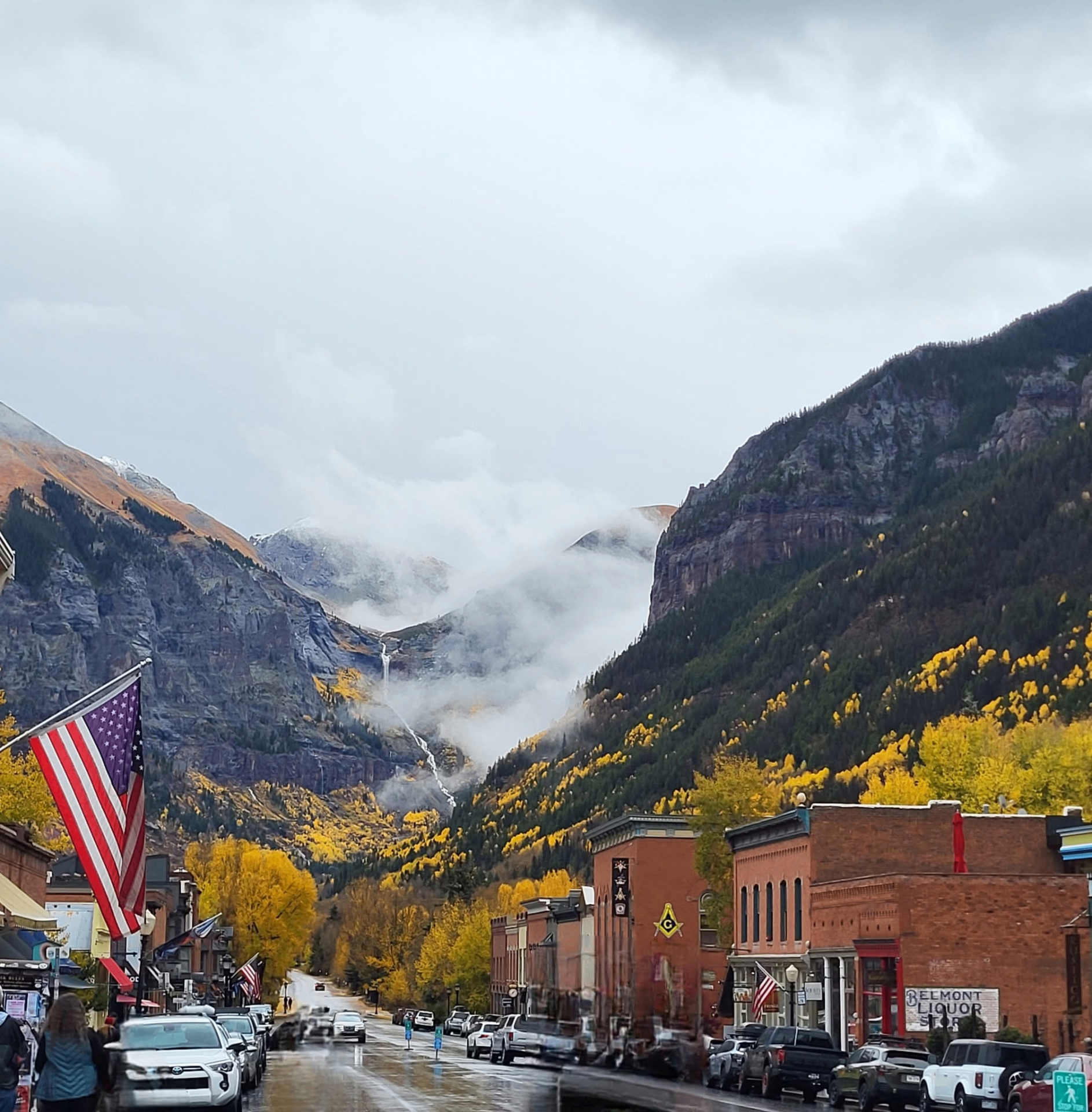 A scenic mountain town main street lined with historic brick buildings and golden autumn trees, with misty peaks rising in the background
